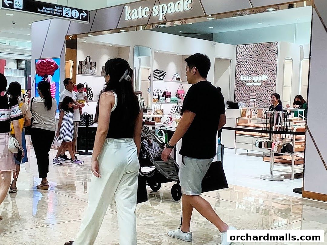 People walk past the Kate Spade New York store entrance displaying handbags and accessories.