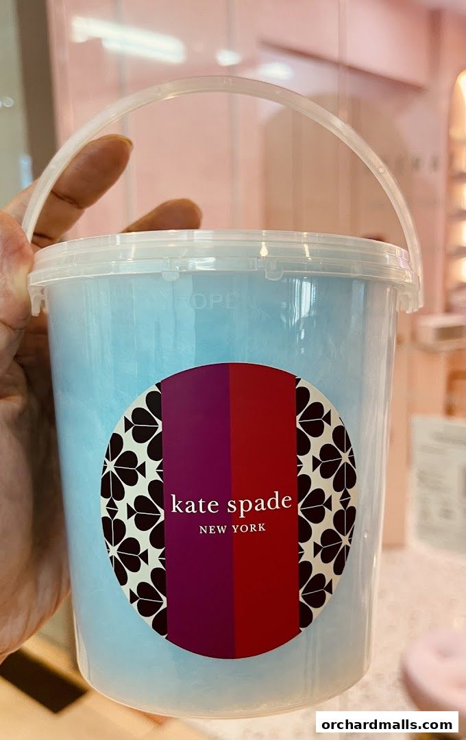 A hand holds a clear bucket filled with blue cotton candy, branded with Kate Spade New York.