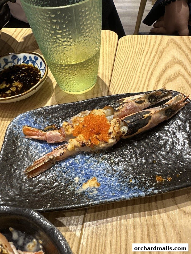 Two grilled prawns topped with roe served on a dark plate with soy sauce and a drink.