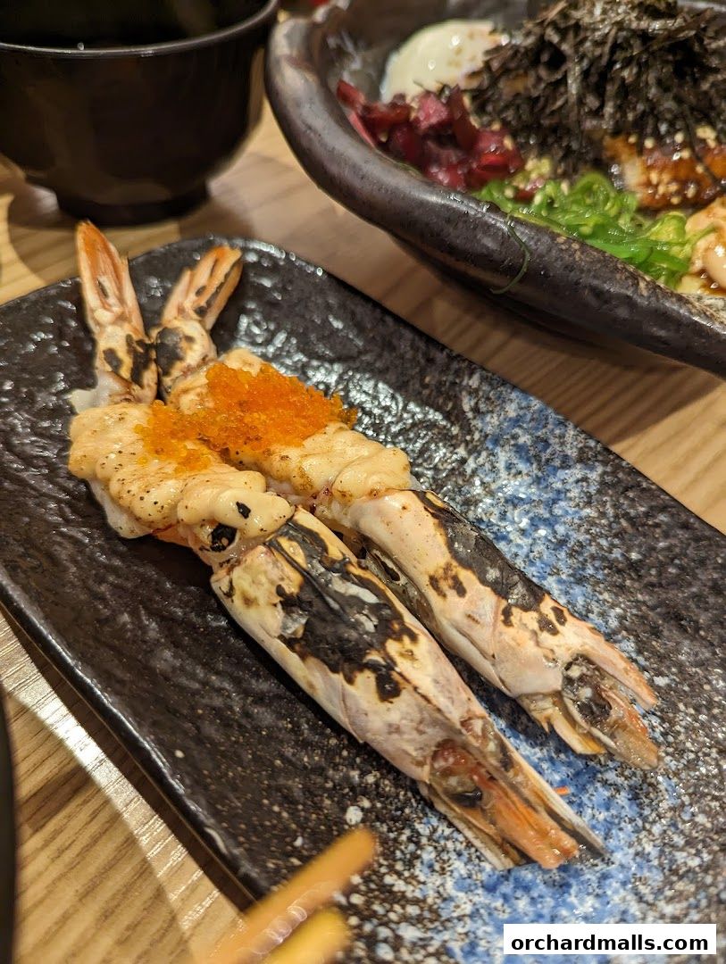 Close-up of grilled shrimp sushi with roe on a dark plate, with another dish in the background.