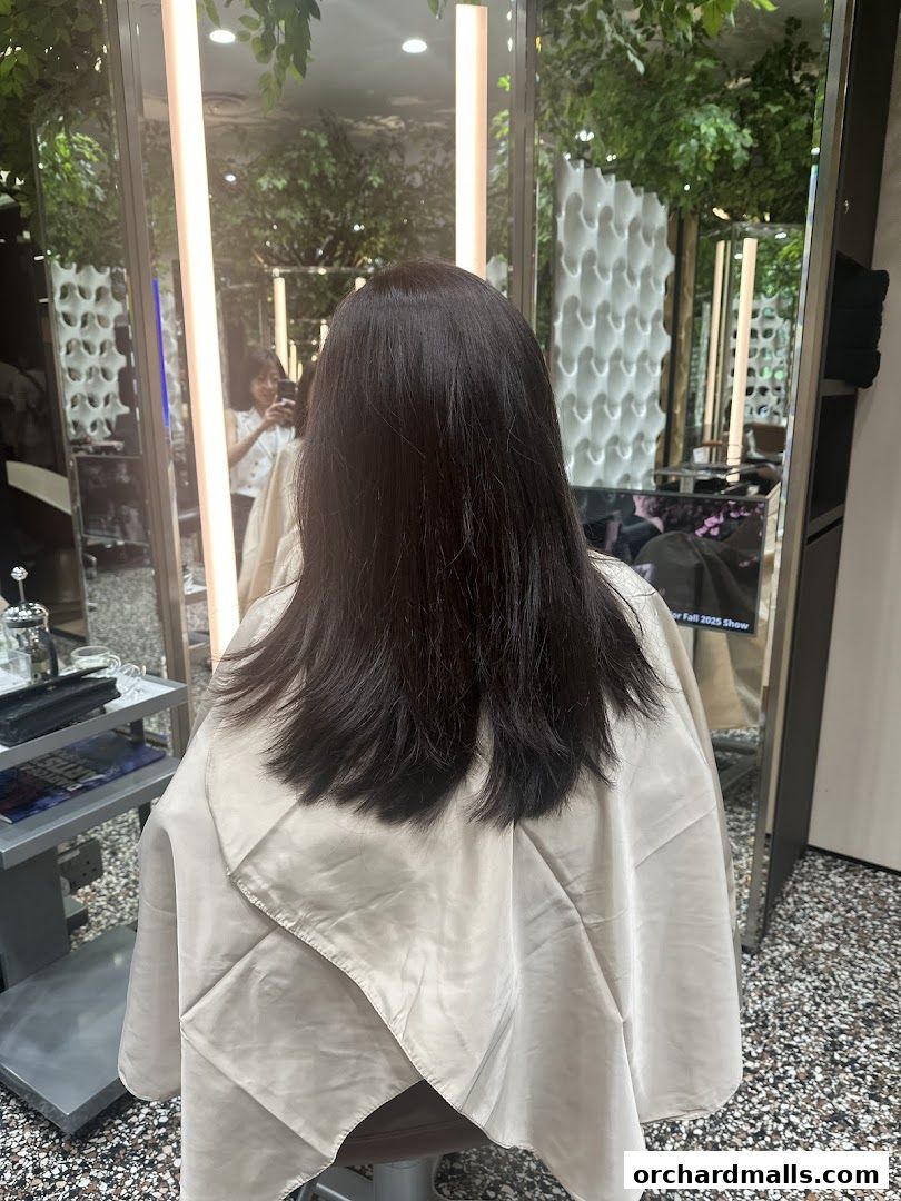 A person with long dark hair sits in a salon chair, draped in a cape.