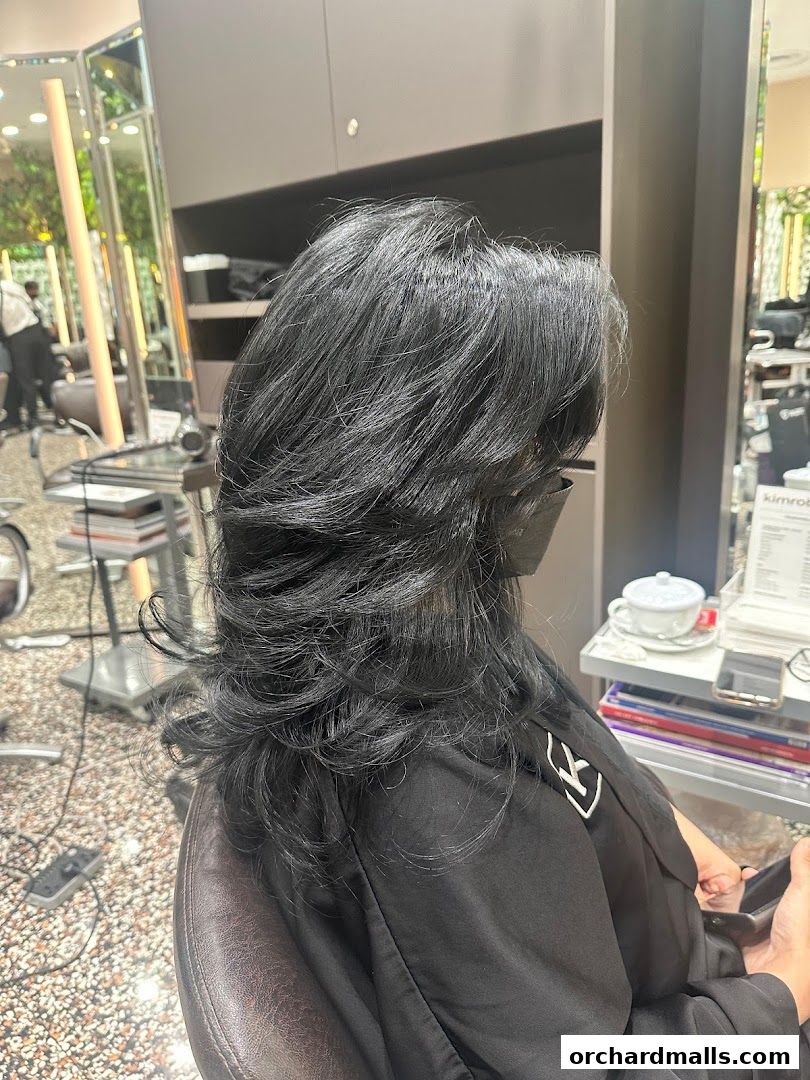A person with long, layered black hair sits in a salon chair, wearing a black cape.