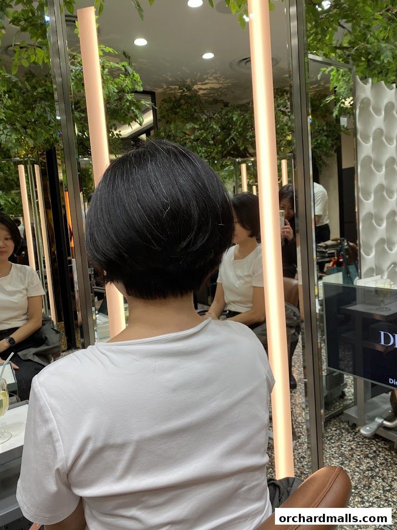 A person with short black hair sits in a salon chair, reflected in mirrors.