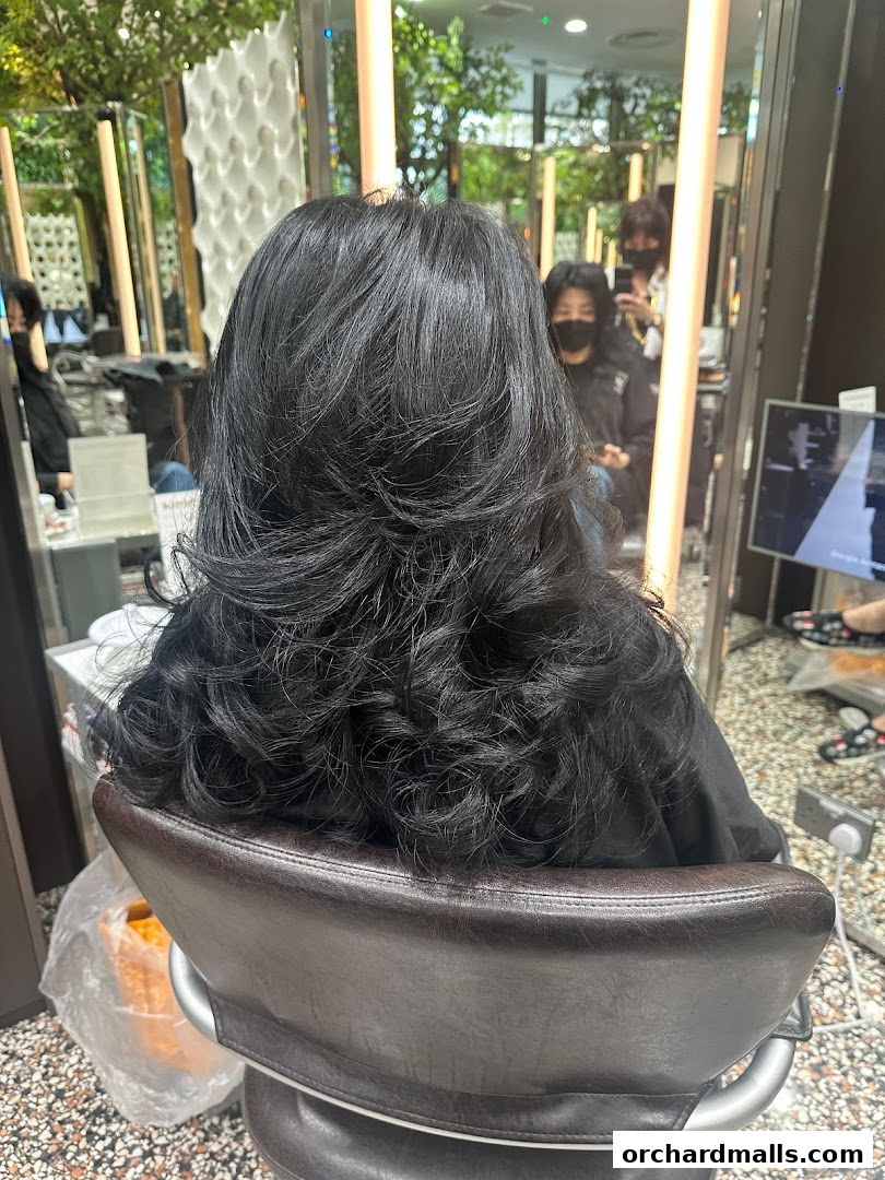 A person with long, layered black hair sits in a salon chair, viewed from behind.