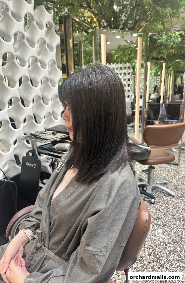 A woman with dark hair sits in a salon chair, wearing a grey robe.
