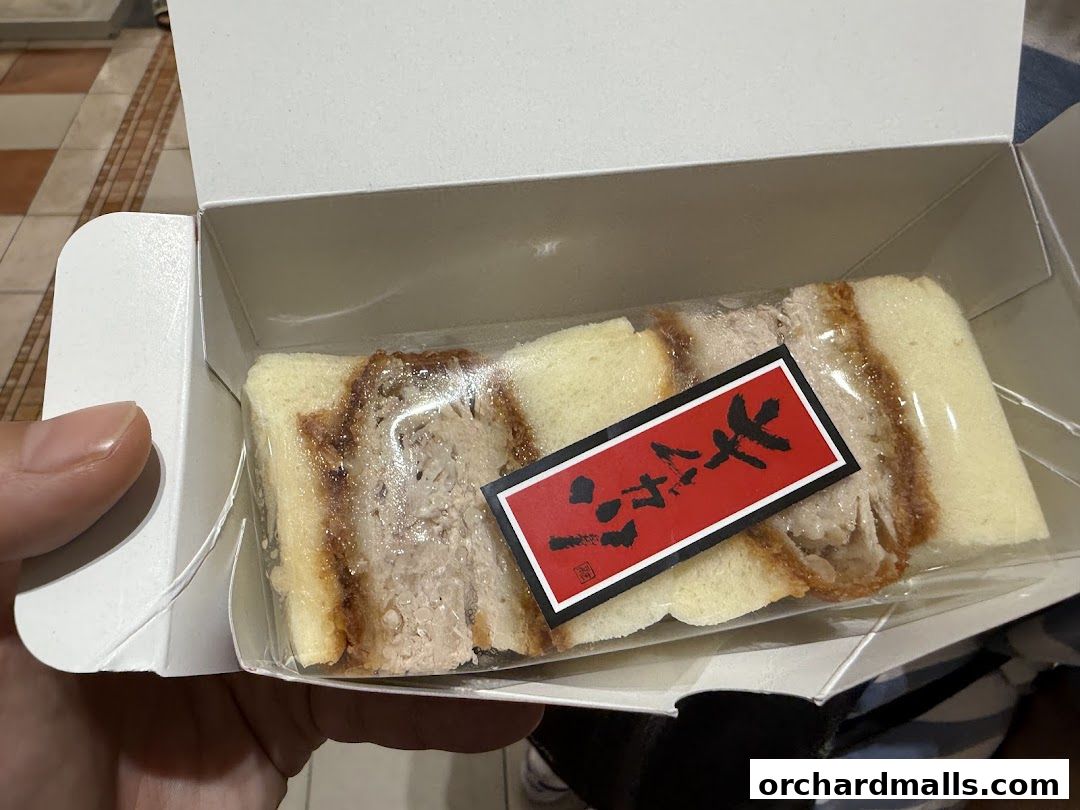 A hand holding a white box containing a katsu sandwich with a red label.