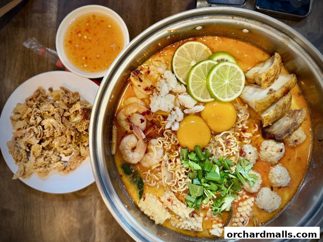 A steaming pot of Tom Yum soup with noodles, shrimp, eggs, and pork belly, served with crispy fried snacks.