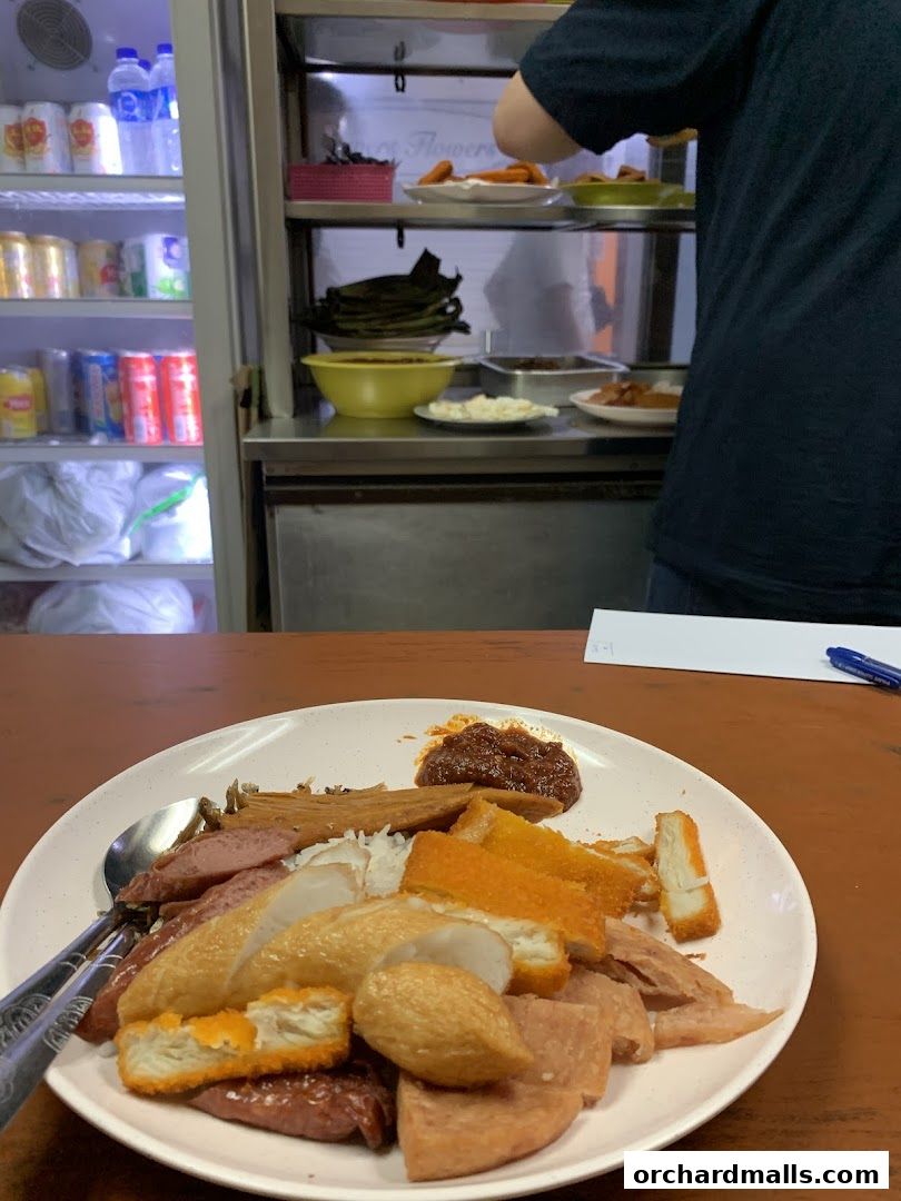 A plate of nasi lemak with various toppings and a person preparing food in the background.