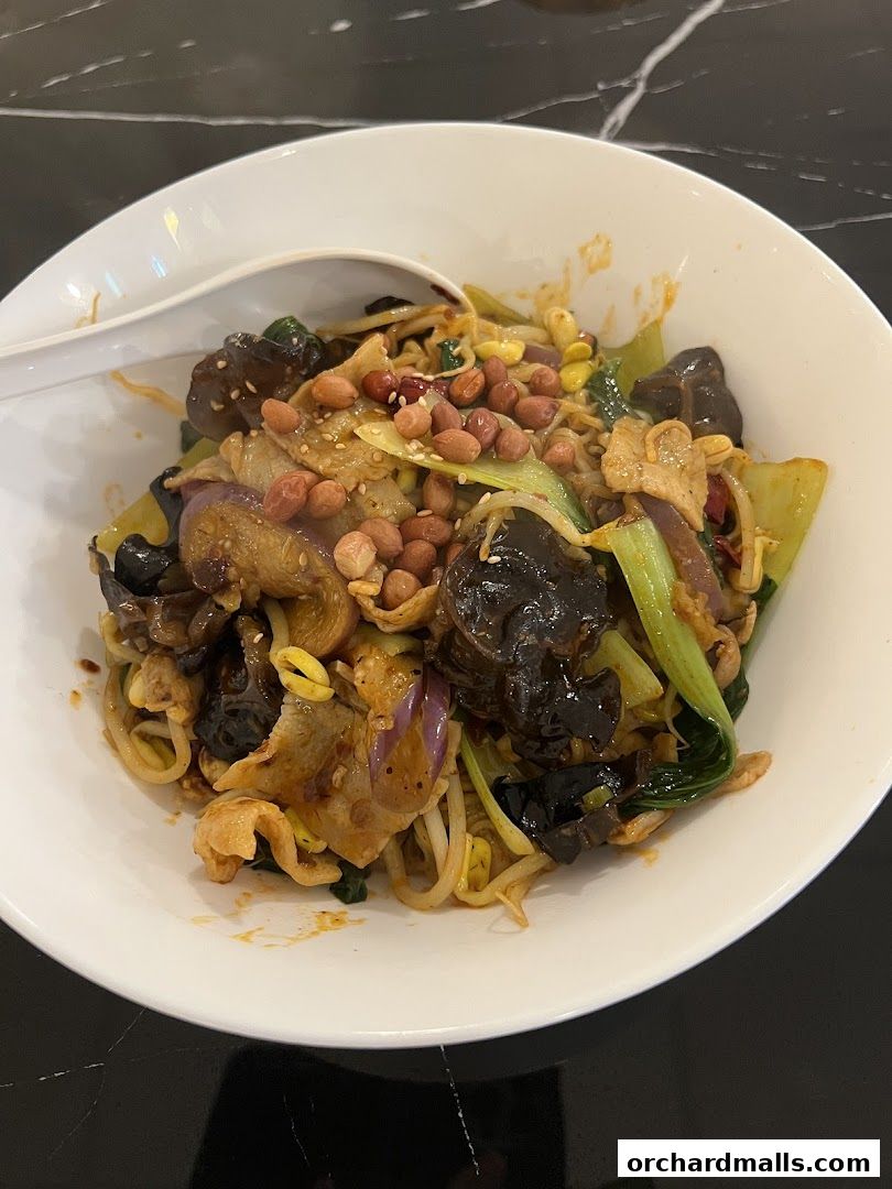 A bowl of spicy noodles with vegetables, peanuts, and meat, served with a spoon.