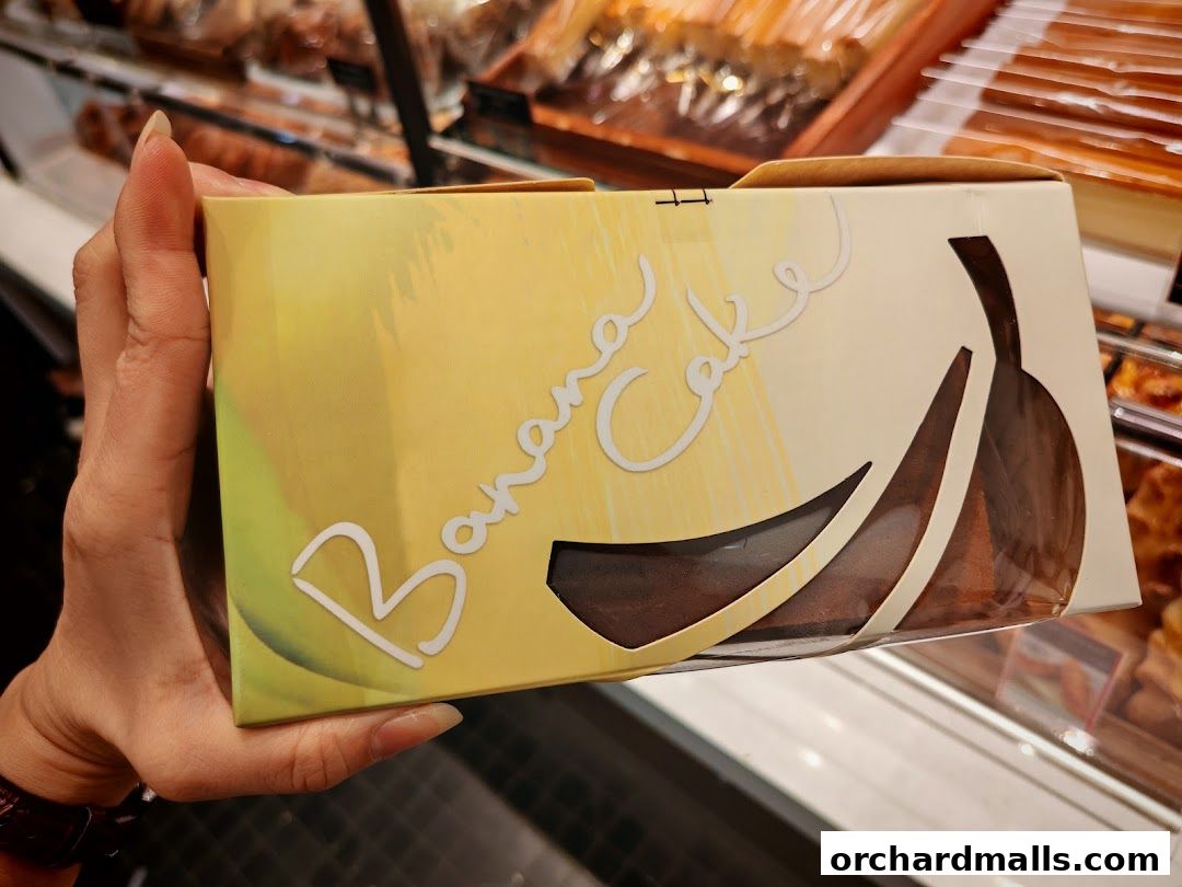 A hand holds a box of banana cake with a window showing the cake inside.