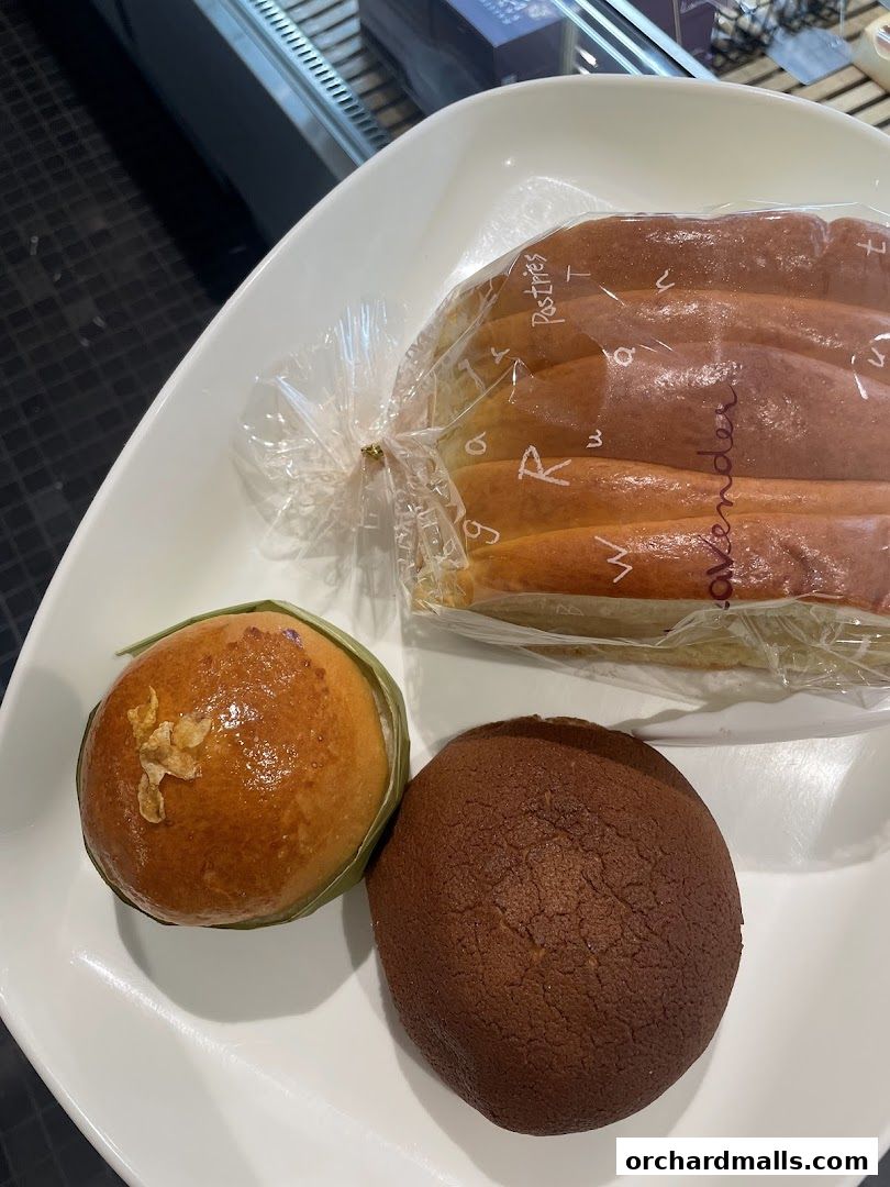 A white plate holds three baked goods: a bun wrapped in plastic, a small round bun with green leaf wrapping, and a dark brown bun.