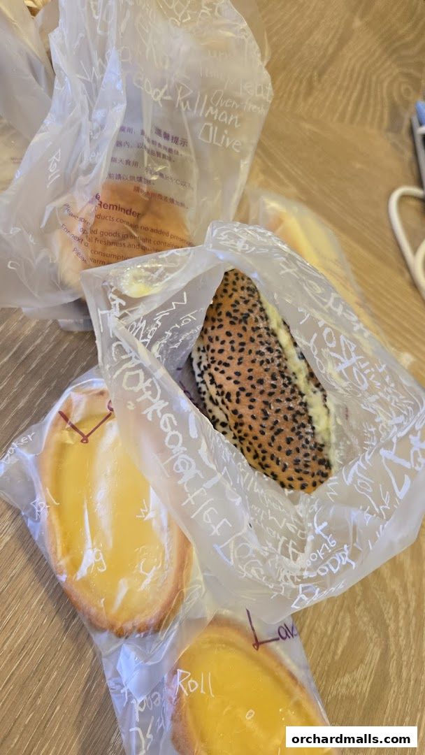 A collection of freshly baked goods, including tarts and a sesame-seed bread roll, packaged in clear plastic bags.
