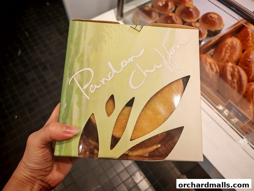A hand holds a box of Pandan Chiffon cake with a bakery display in the background.