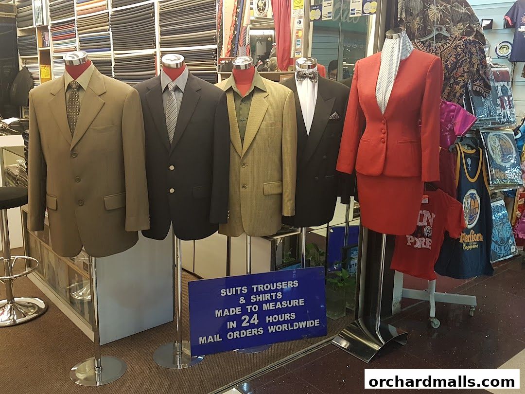 Mannequins display suits and a skirt suit outside a tailor shop with fabric rolls in the background.
