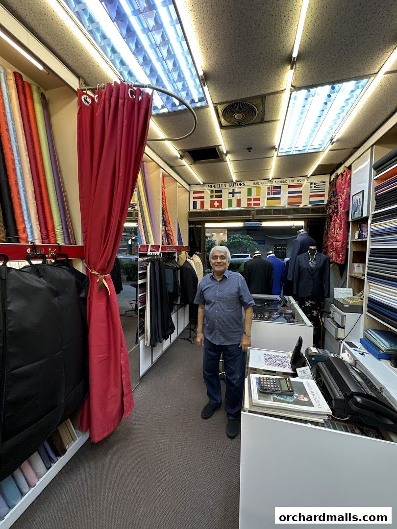 A tailor stands inside his shop, surrounded by fabric rolls and finished garments.