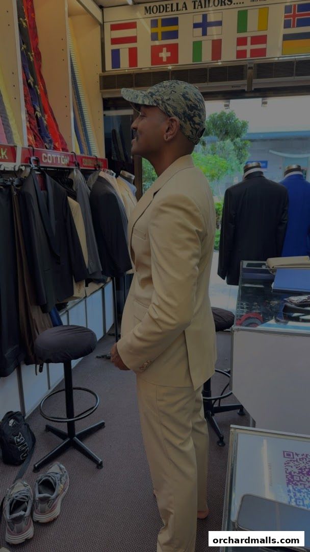 A man in a tan suit stands in a tailor shop, looking at fabric samples.