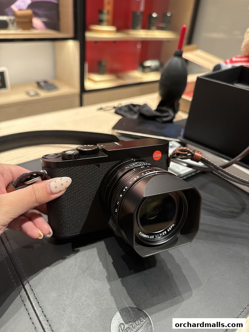A hand holds a black Leica camera with a lens attached, on a wooden counter.