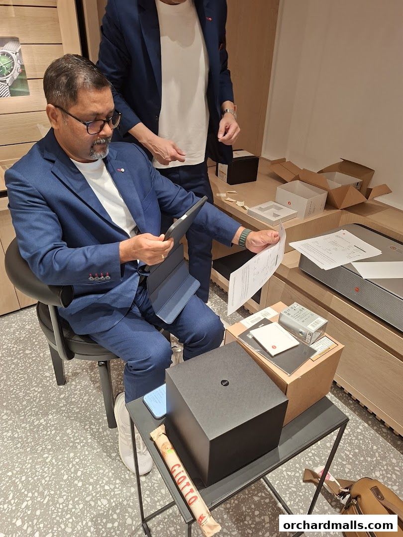 A man in a blue suit sits with a tablet and papers, with another person standing behind him.