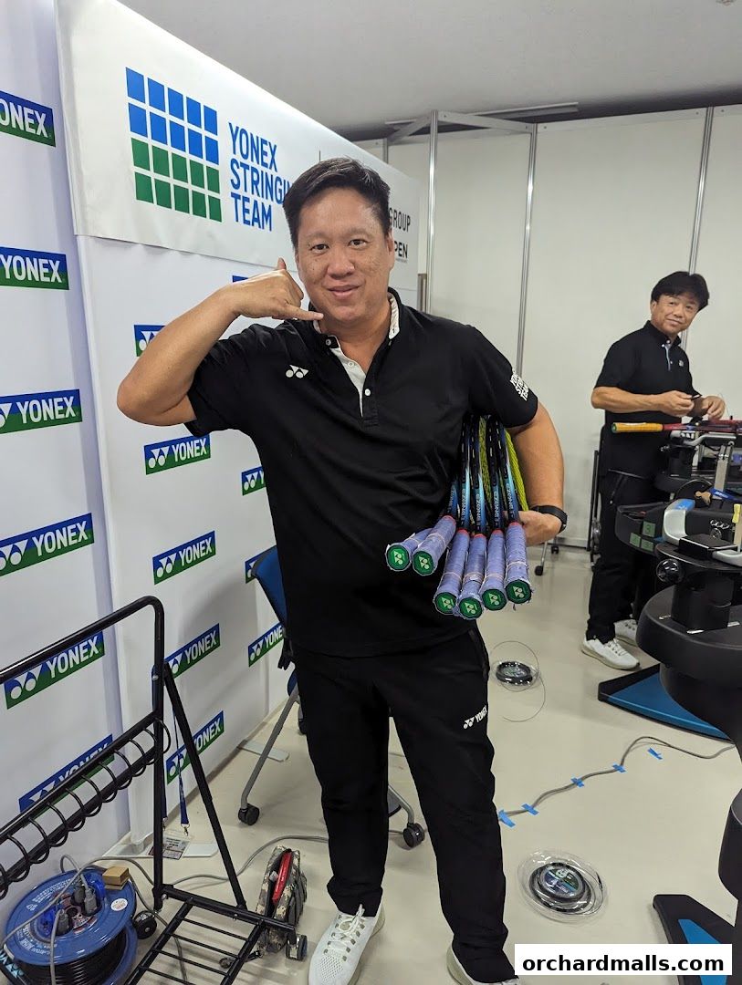 A man holds several badminton rackets while making a call gesture. Another man strings rackets in the background.