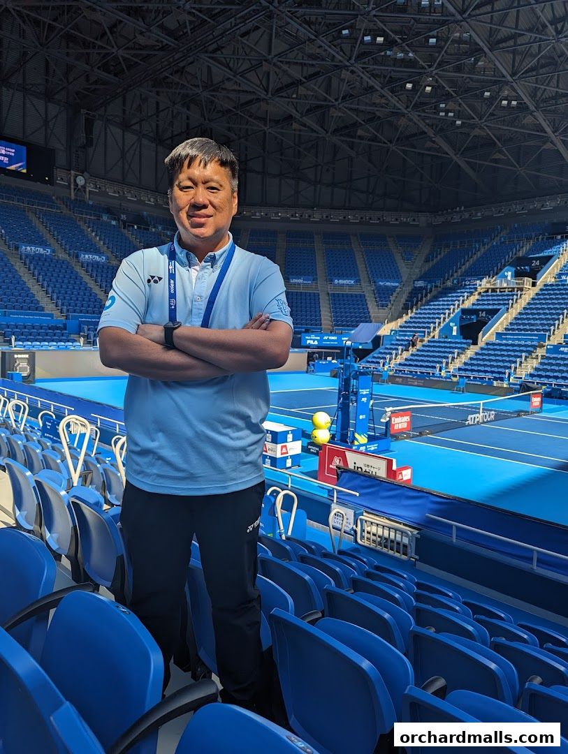 Man in sportswear stands in a stadium with blue seats and a tennis court.