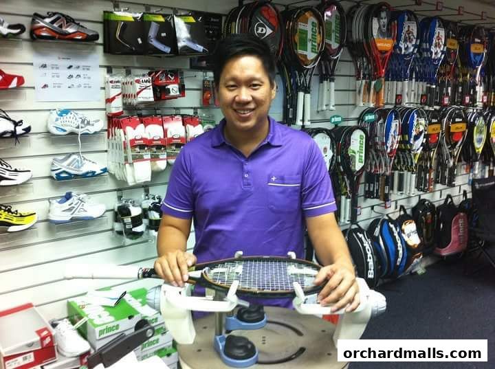 A man in a purple shirt stands in a sports shop with tennis rackets and shoes on display.