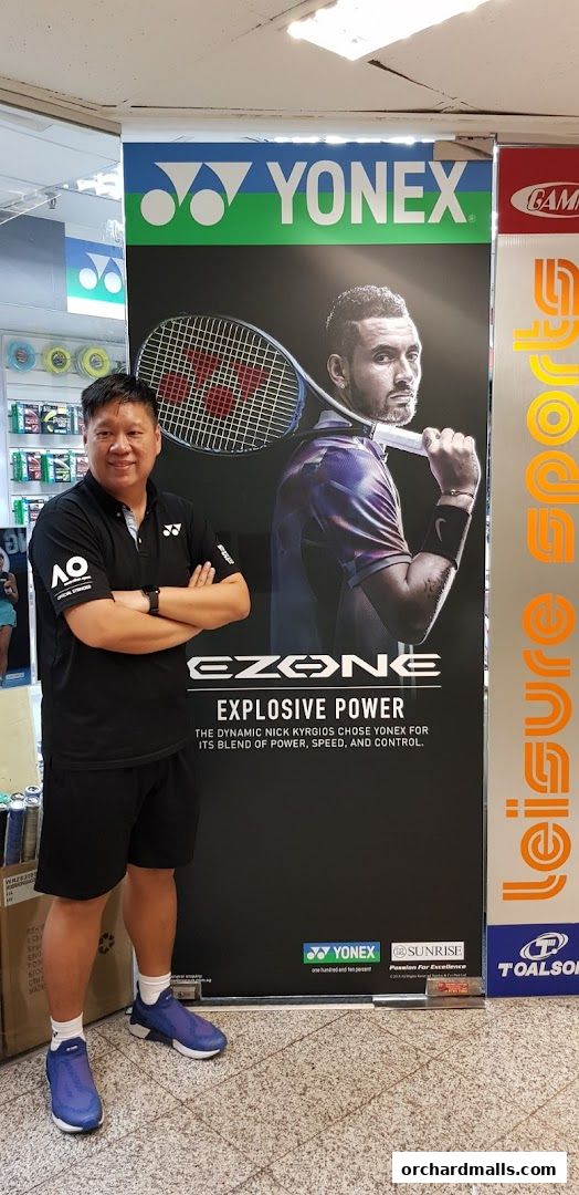 A man stands in front of a large Yonex banner featuring Nick Kyrgios and the Ezone racket.