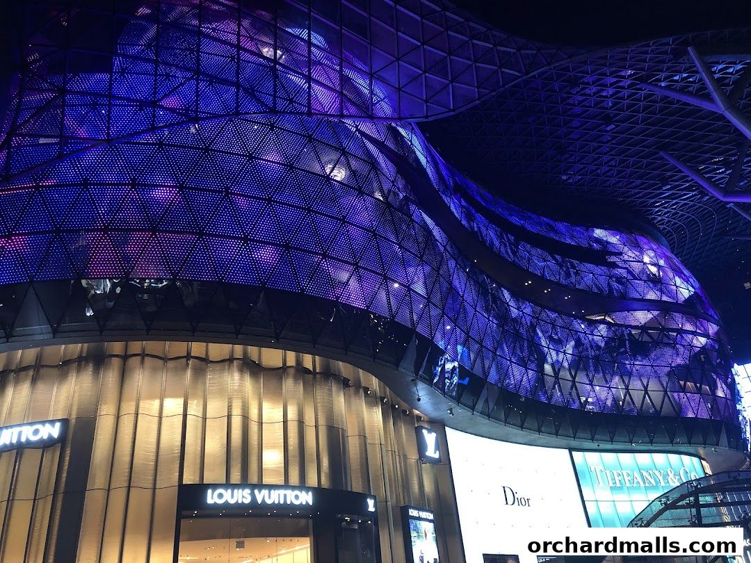 The exterior of a modern building at night, illuminated with purple lights and featuring prominent brand names.