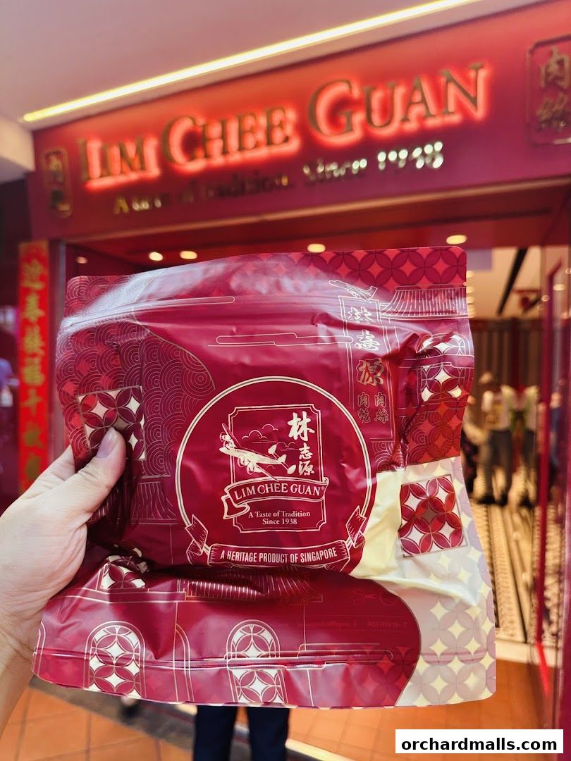 A hand holds a red package with the Lim Chee Guan logo in front of the store.