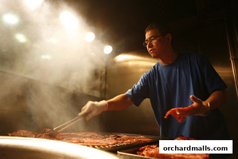 A man in a blue t-shirt grills meat with tongs, surrounded by smoke.