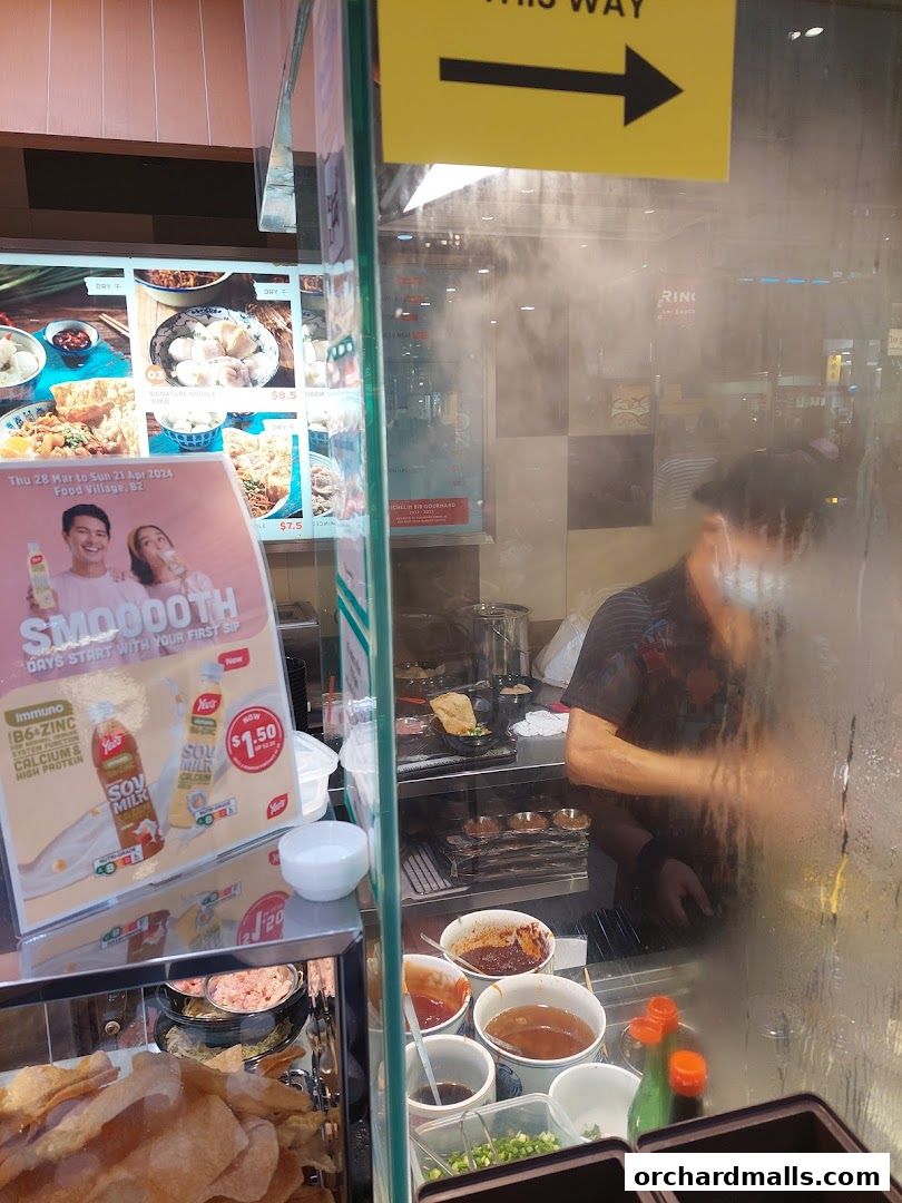 A food stall with various ingredients and a cook preparing food behind a glass partition.