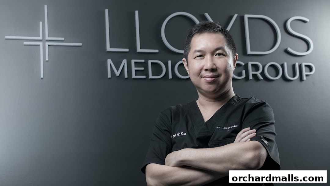 A doctor stands in front of the Lloyds Medical Group logo with arms crossed.
