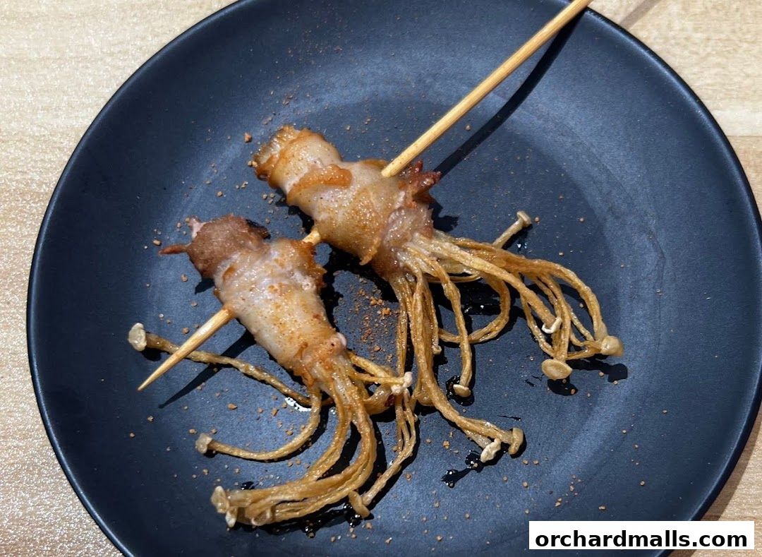 Two skewers of enoki mushrooms wrapped in bacon on a black plate.