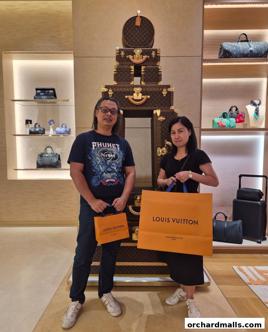A couple poses with shopping bags in front of a Louis Vuitton store display.