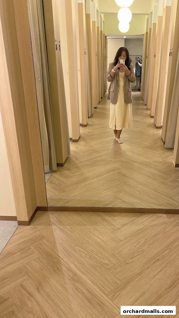 A woman takes a mirror selfie in a fitting room with beige curtains and wooden floors.