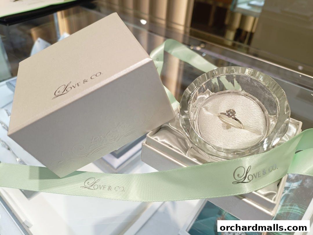 A diamond engagement ring displayed in a crystal holder with a Love & Co. gift box and ribbon.