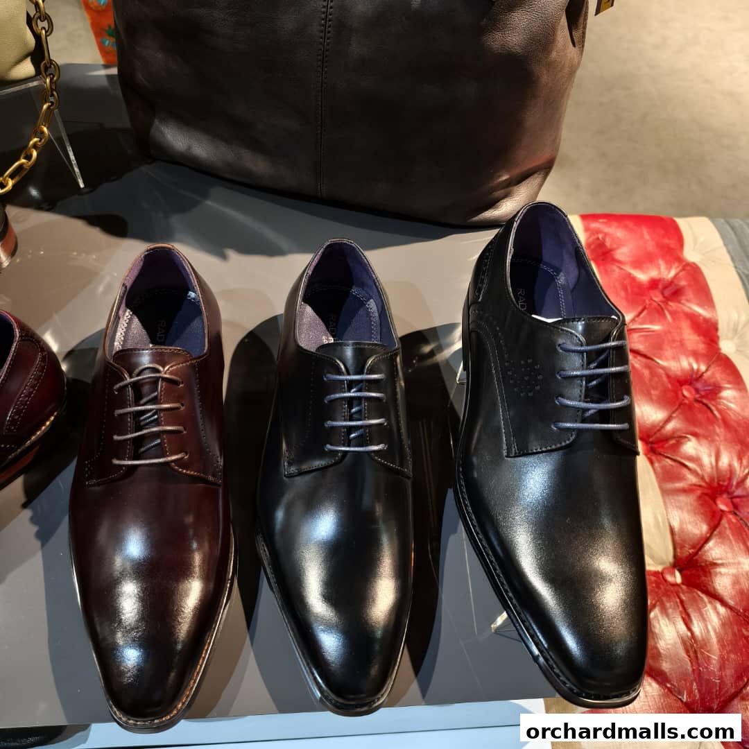 Three pairs of men's dress shoes are displayed on a surface with handbags in the background.