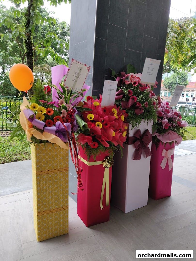 A display of congratulatory flower stands with an orange balloon outside a building.