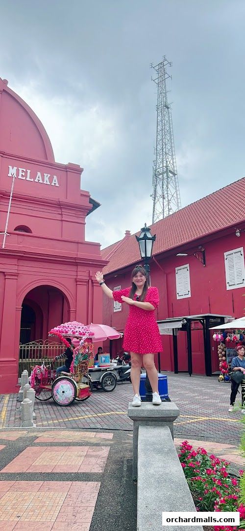 A woman poses in front of a pink building with a decorated rickshaw and a tall tower.