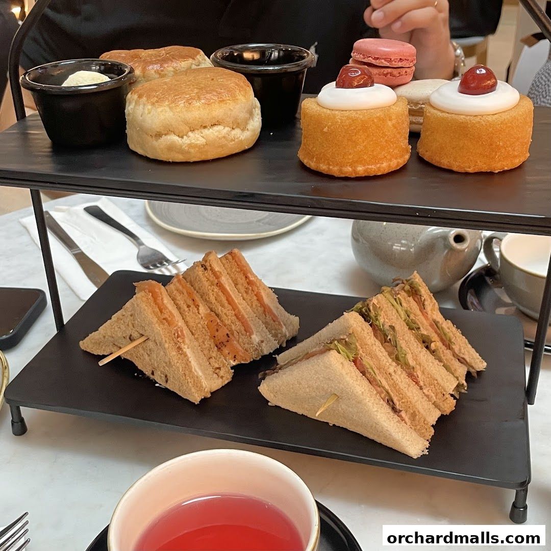 A tiered stand displays sandwiches, scones, mini cakes, and macarons, with tea in a cup.