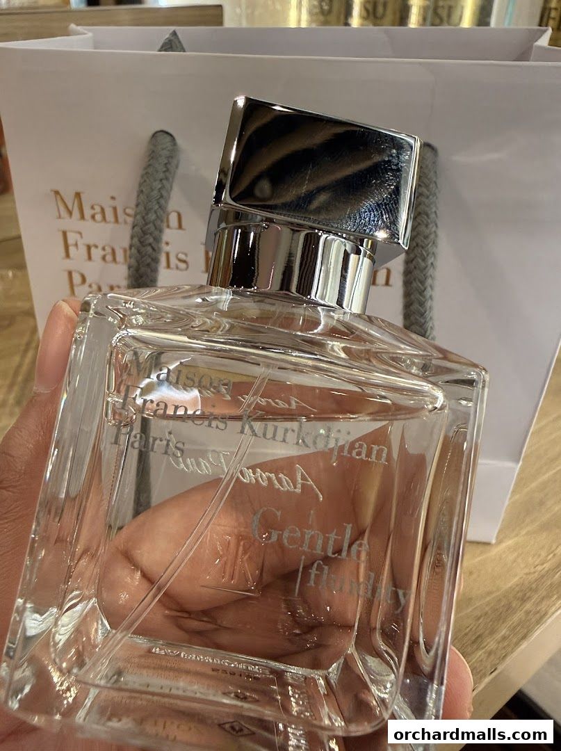 A hand holds a clear glass perfume bottle with a silver cap, in front of a branded shopping bag.
