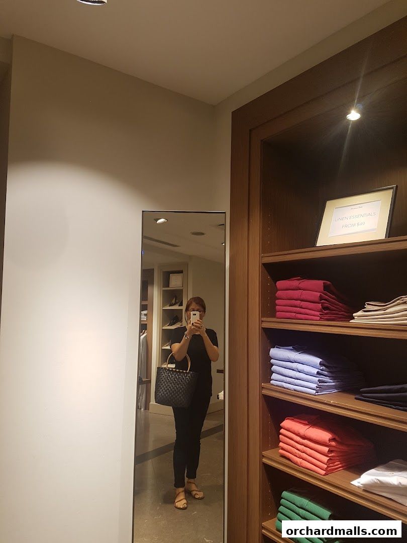 A woman takes a mirror selfie in a clothing store, showcasing folded shirts on shelves.