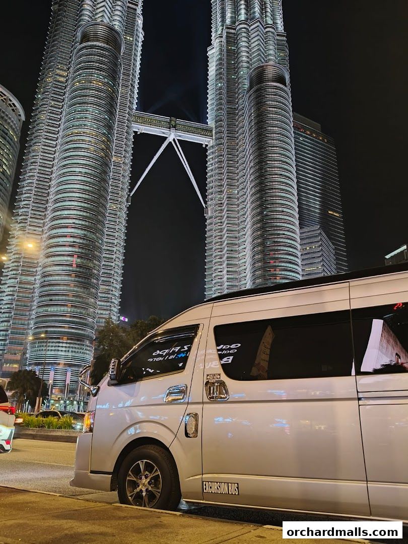 A silver excursion bus parked on the street with the Petronas Twin Towers in the background at night.