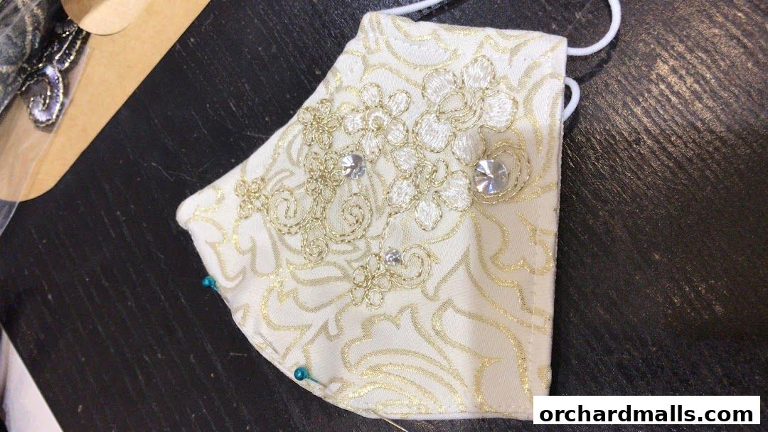 A close-up of an ornate, white and gold embroidered face mask with rhinestones.