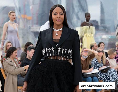 Naomi Campbell walks a runway in a black blazer and feathered skirt, with models and audience behind her.