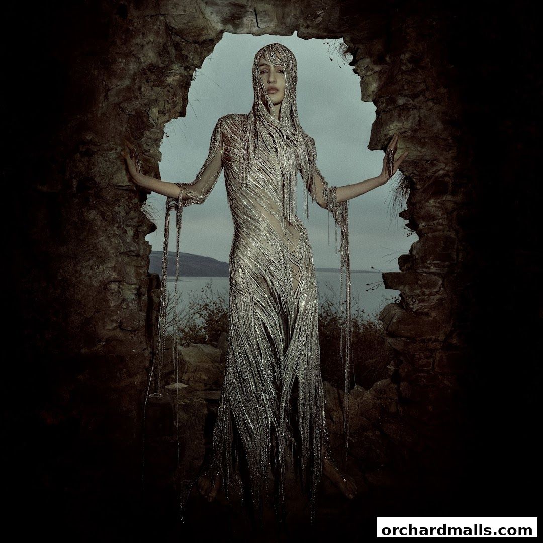 A model wears a shimmering, fringed gown and headpiece, posing in a stone archway.