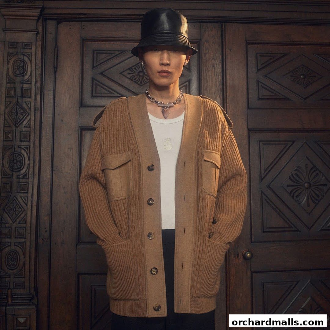 A person wearing a brown cardigan, white t-shirt, and black hat stands in front of a wooden door.