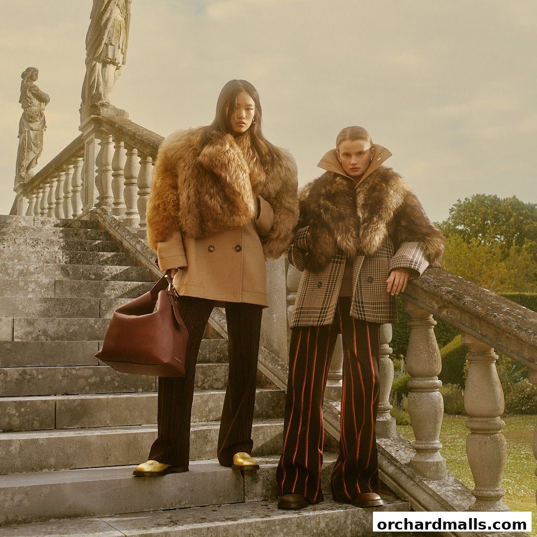 Two models pose on a stone staircase wearing luxurious fur-trimmed coats and stylish trousers.