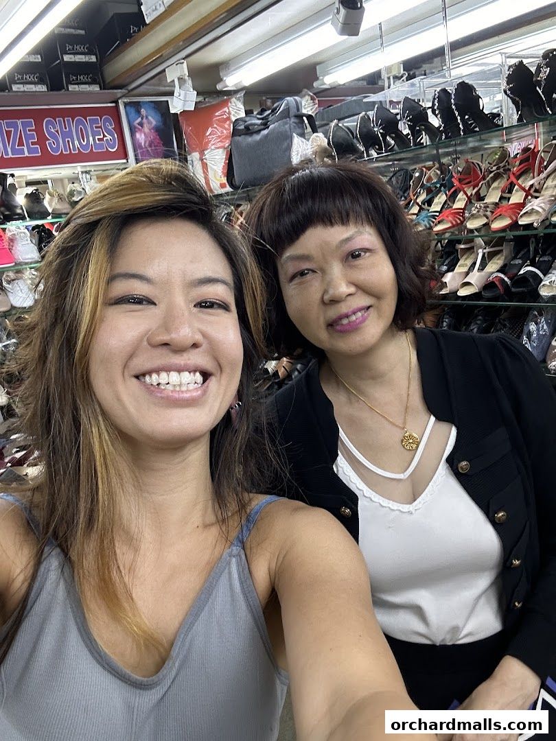 Two smiling women pose in a shoe boutique with shelves of shoes in the background.