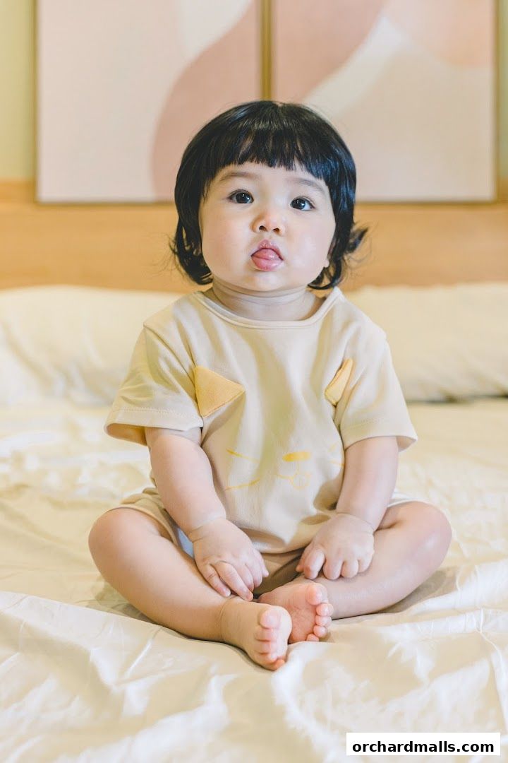 A cute baby with black hair and a light-colored outfit sits on a bed.