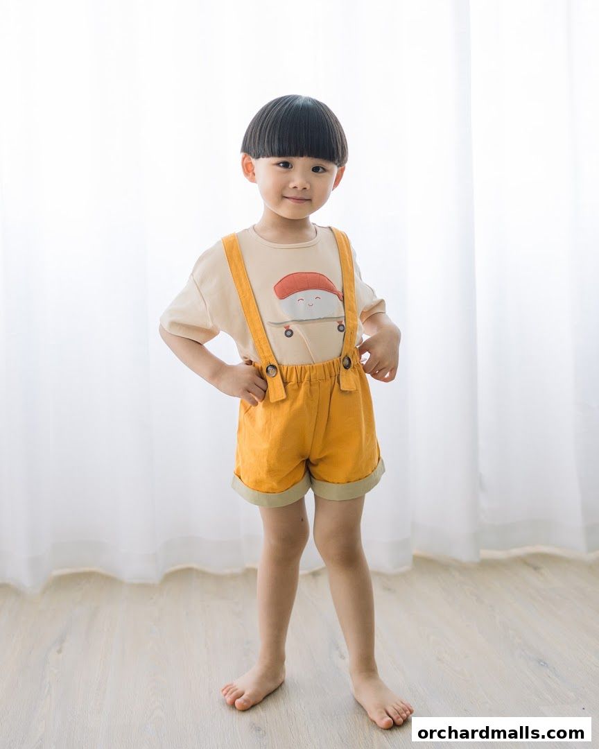 A young child poses in a cute outfit featuring a sushi-themed t-shirt and yellow overalls.